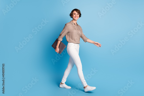 Full length profile portrait of pretty cheerful lady walking look camera hold netbook isolated on blue color background