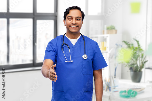 healthcare, profession and medicine concept - happy smiling indian doctor or male nurse in blue uniform with stethoscope giving hand for handshake over medical office at hospital on background