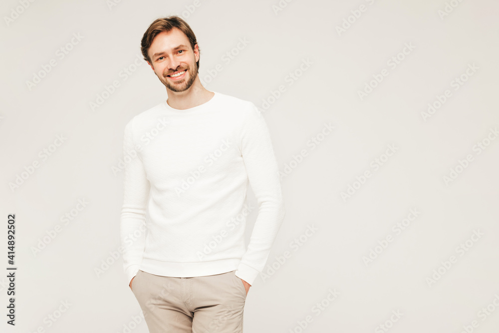 Portrait of handsome smiling hipster lumbersexual businessman model wearing casual white sweater and trousers. Fashion stylish man posing against gray background in studio
