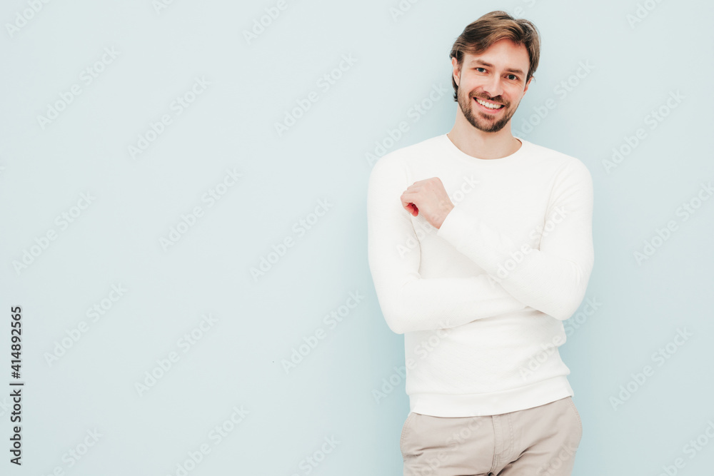 Obraz premium Portrait of handsome smiling hipster lumbersexual businessman model wearing casual white sweater and trousers. Fashion stylish man posing against light blue wall in studio