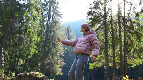 Young adult caucasian beautiful woman in warm jacket and wool suit video chating by cell phone to friends or family during walk in scenic green woods pine forest.Person make selfie walking outdoors