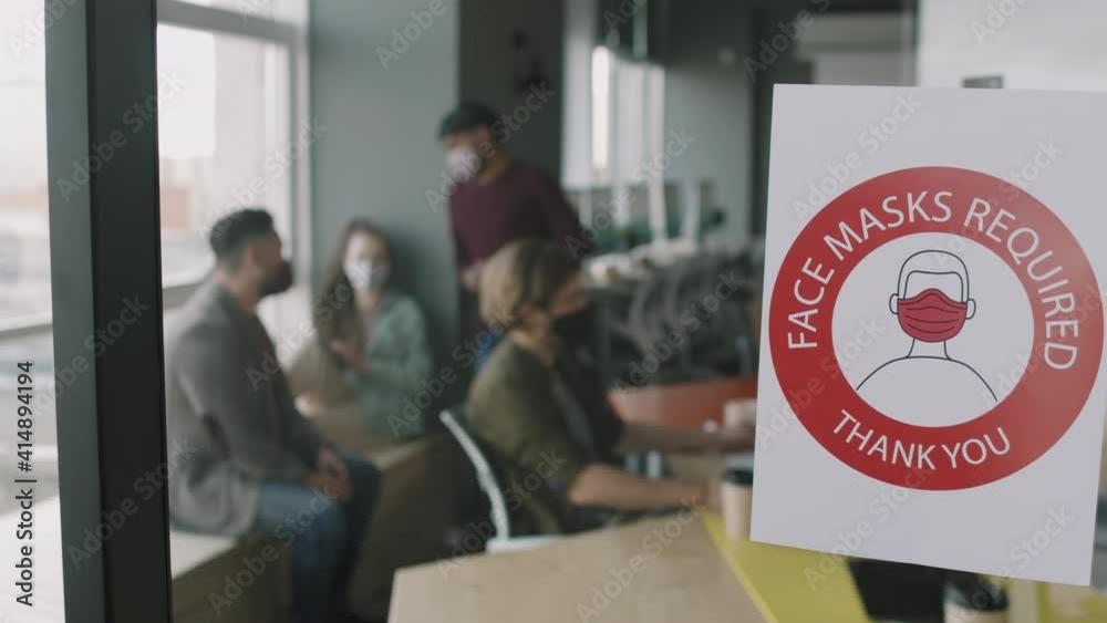 PAN slowmo of Face Masks Required sign on glass wall in office. Businesspeople in face masks working inside PAN slowmo of Face Masks Required sign on glass wall in office. Businesspeople in face masks working inside
