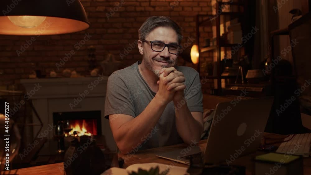 Home office - older white man working from home. Thinking, typing on laptop computer. Sitting at desk in dark living room iin front of fireplace.