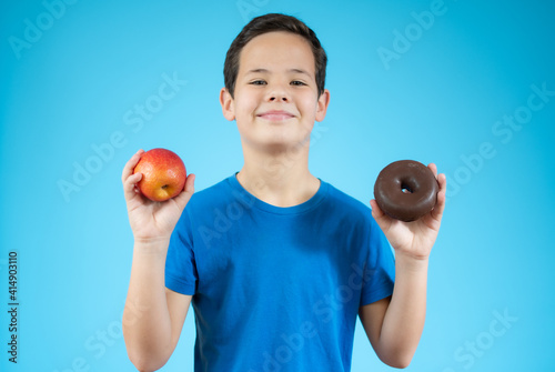 Young boy choosing between an apple and donuts isolated over blue background photo