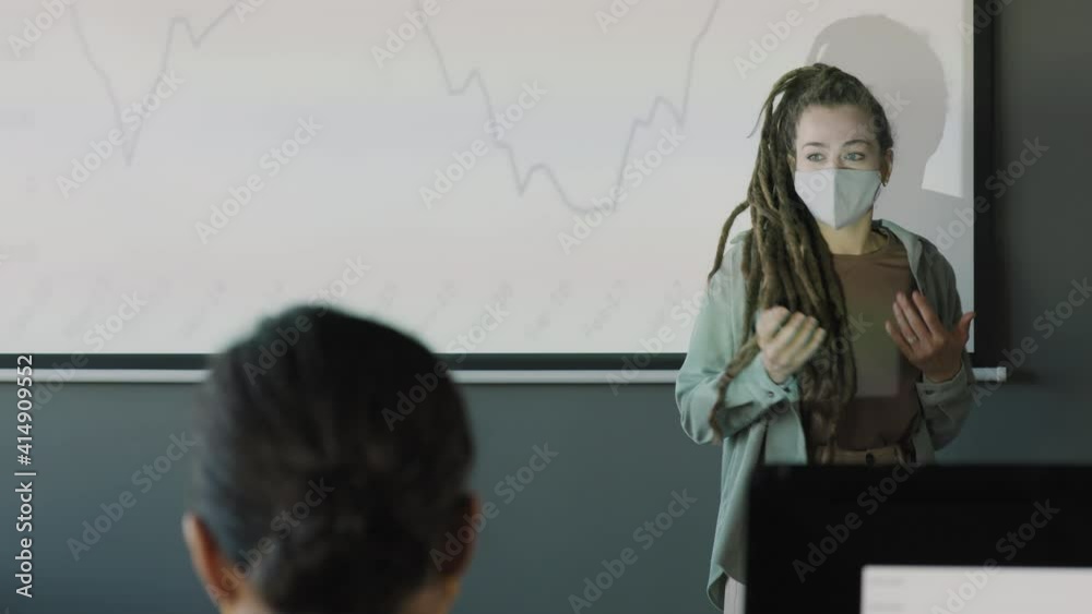Handheld tracking of young businesswoman in face mask walking before projector screen with line chart and delivering financial report to unrecognizable colleagues Handheld tracking of young businesswoman in face mask walking before projector screen with line chart and delivering financial report to unrecognizable colleagues