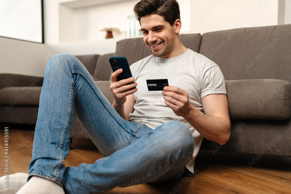 Fototapeta premium Happy guy using mobile phone and credit card while sitting on floor