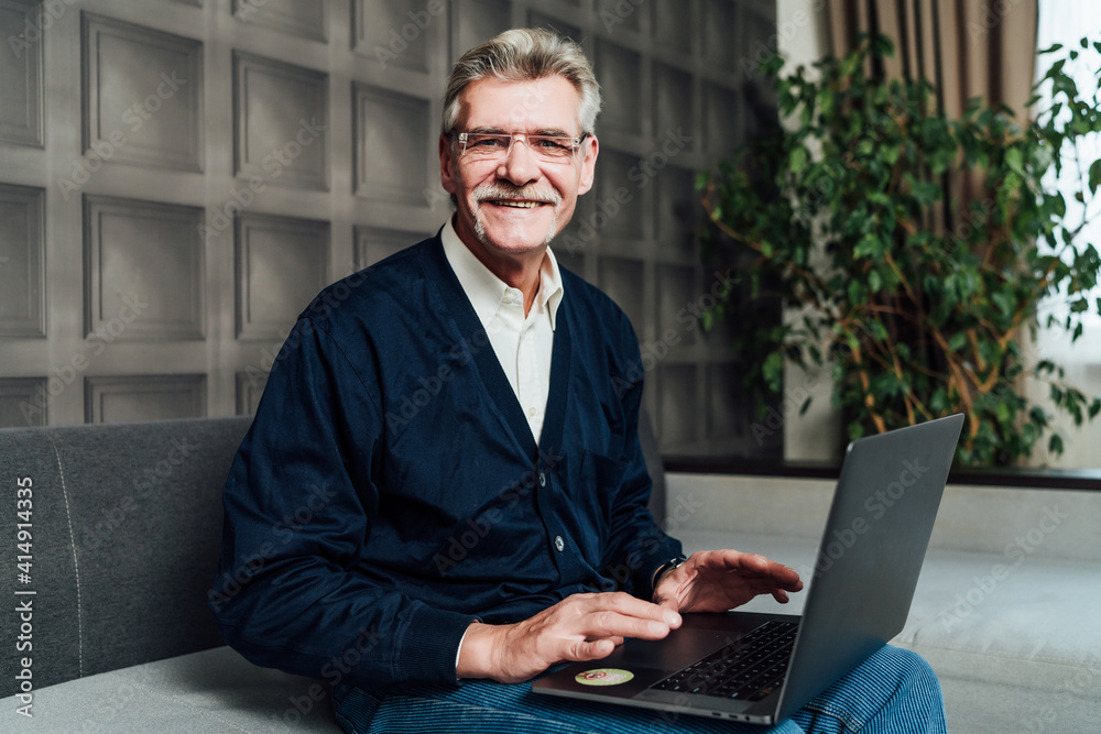 Portrait of a cheerful old grandfather sitting on a sofa with a computer he looks at the camera and smiles Portrait of a cheerful old grandfather sitting on a sofa with a computer he looks at the camera and smiles