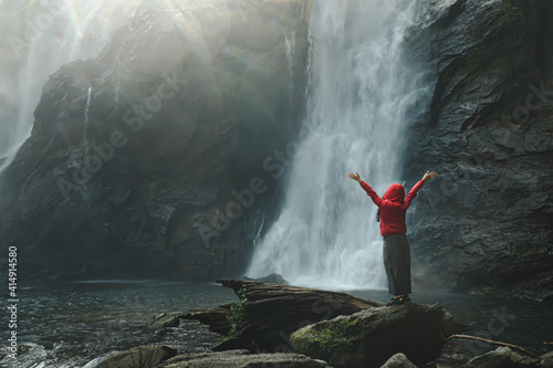 Picture from the back of a woman in a red hood raise both arm and standing on rock in waterfall. Large amounts of water flowed strongly, and scattering droplets. Idea for traveling background. © Pang wrp