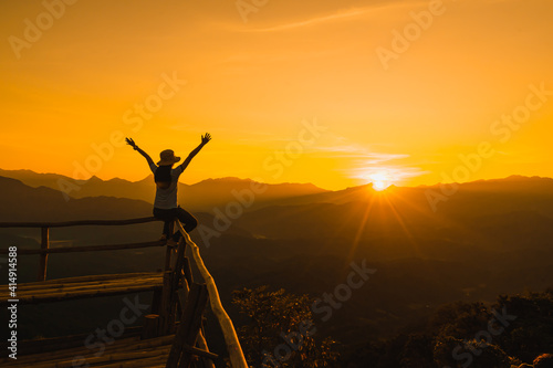 Picture from the back of a woman sitting and raise both arm on a wooden porch extending into a high mountain cliff. The sun is setting on the mountain and there is a beautiful warm orange light. © Pang wrp