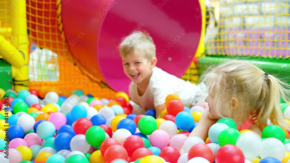 Having fun in playroom. Children boy and girl playing on indoor playground in multi colored plastic balls in big dry paddling pool in playing centre down slide and swing. Leisure Activity.