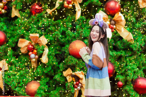 A beautiful Asian woman in a colorful sweater stands smiling and happy in front of the Christmas tree With bokeh as a background in the theme to celebrate Christmas and Happy New Year