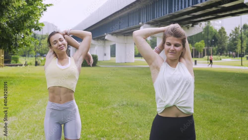 Athletic women in sportswear do exercises in modern city park. Close-up girlfriends standing on lush green grass near railway overpass warmup before workout and stretching arms in summer morning Athletic women in sportswear do exercises in modern city park. Close-up girlfriends standing on lush green grass near railway overpass warmup before workout and stretching arms in summer morning