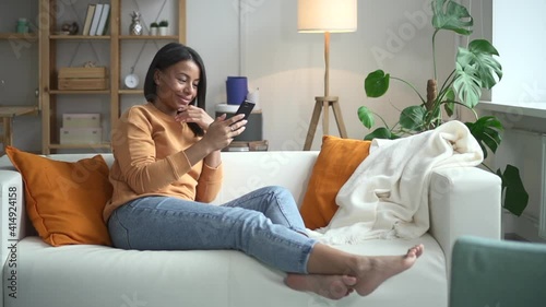 Wallpaper Mural Woman using phone and smiling, sitting on couch in light room spbd. Young American female holds smartphone in hands and looks at screen with happy smile, sits on sofa in home interior. Millennial lady Torontodigital.ca