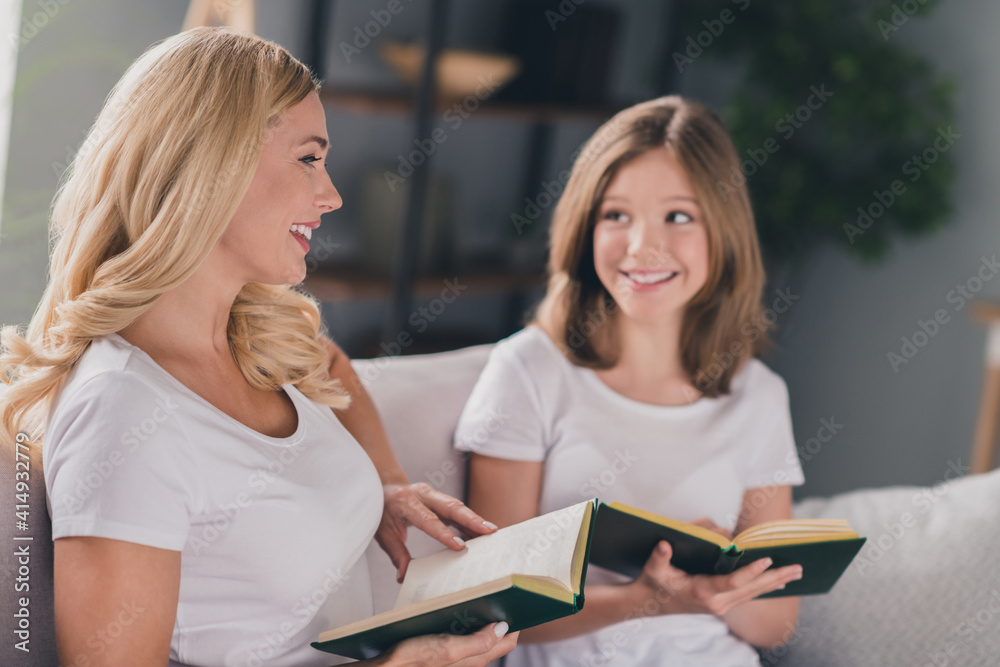 Photo of pretty funny daughter mom wear white t-shirts smiling reading novels sitting sofa indoors inside room home Photo of pretty funny daughter mom wear white t-shirts smiling reading novels sitting sofa indoors inside room home