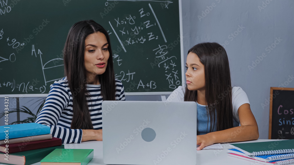 mother and daughter doing homework near laptop and notebooks on table mother and daughter doing homework near laptop and notebooks on table