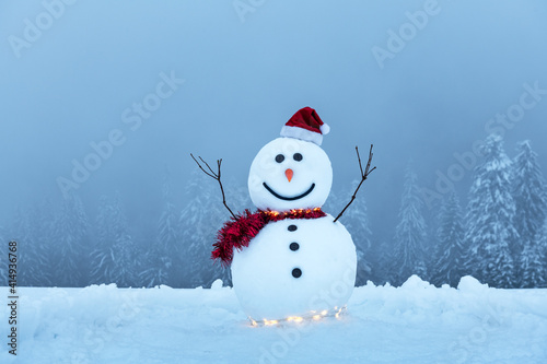 Funny snowman in stylish red hat and red scalf on snowy mountains. Glowing garland for Christmas vibe © Ivan Kmit