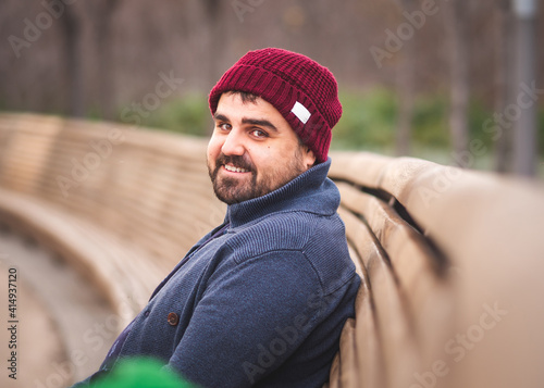 Boarded guy in a woolen hat and sweater sitting on park bench. Looking at the camera and smiling.
