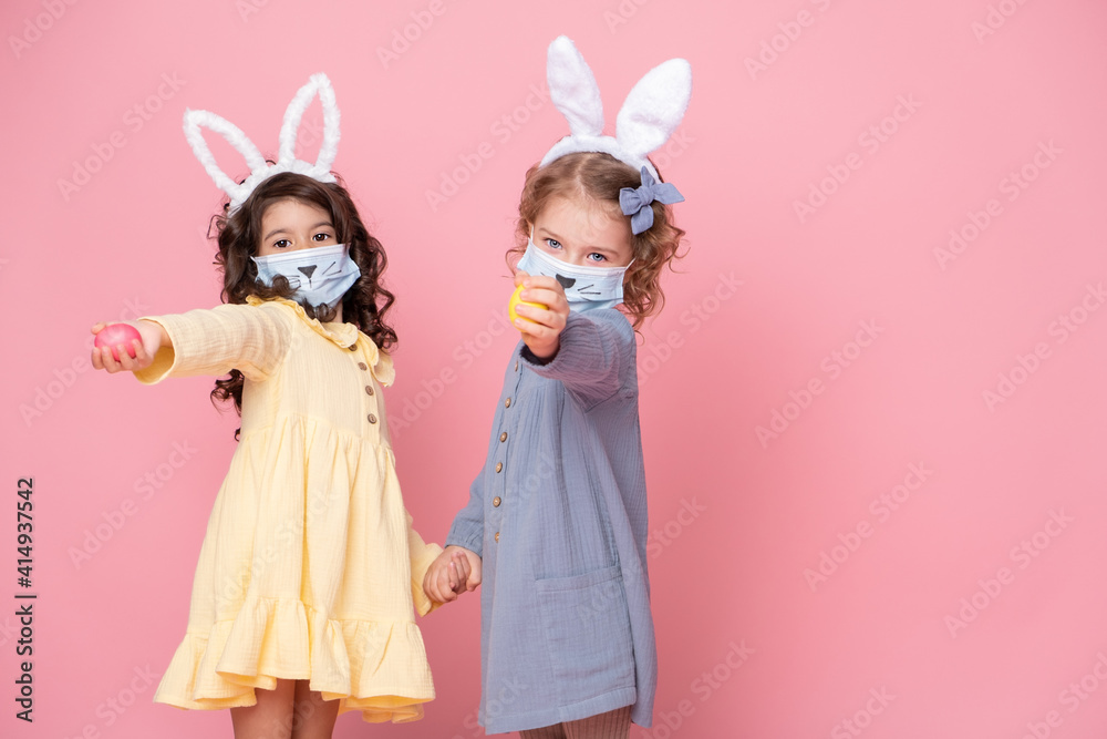 two girls in bunny ears headband and protective mask with colored eggs on pink background. Covid easter concept