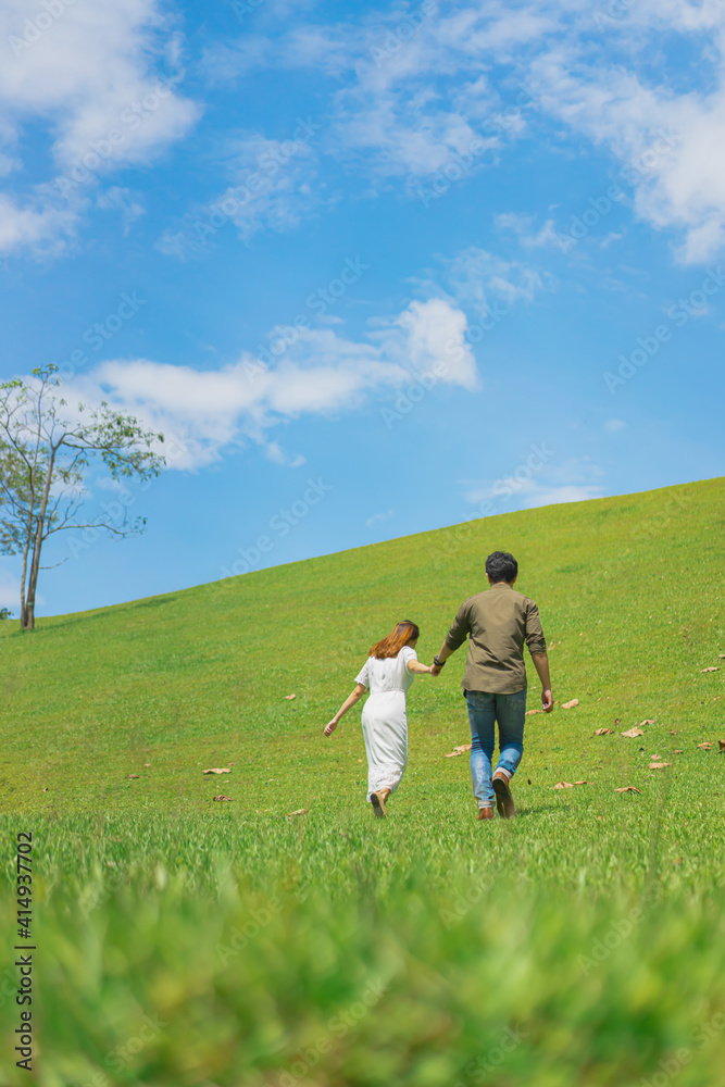 A photo from the back of a man and woman holding hands on a green grassy slope in soft tone. They are walking to the top of the hill on a clear blue sky with white clouds. There is copy space on top.