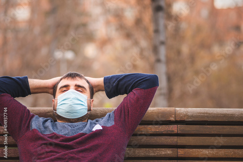 Guy with protective mask relaxing sitting on a park bench at sunset. 