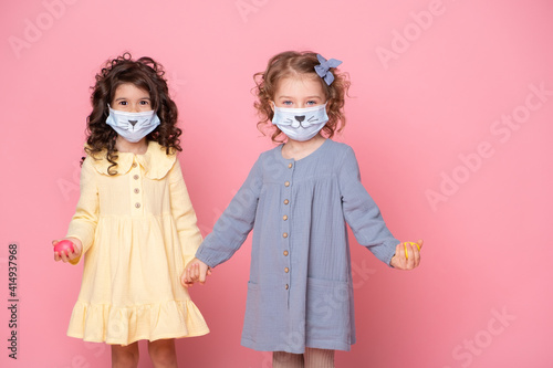 two girls in protective mask with colored eggs holding hands on pink background. Covid easter concept
