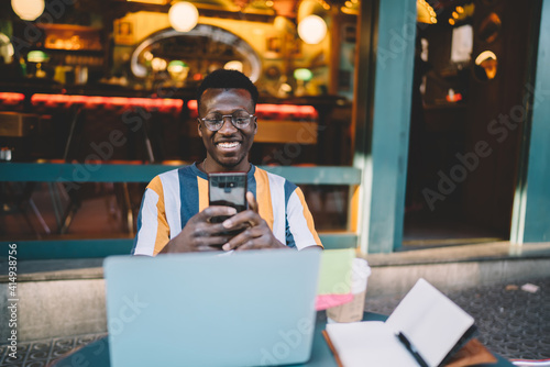 Cheerful male blogger with digital smartphone enjoying networking sitting in street cafe and smiling, toothy hipster guy in optical eyewear using mobile phone for typing text message and sending © BullRun