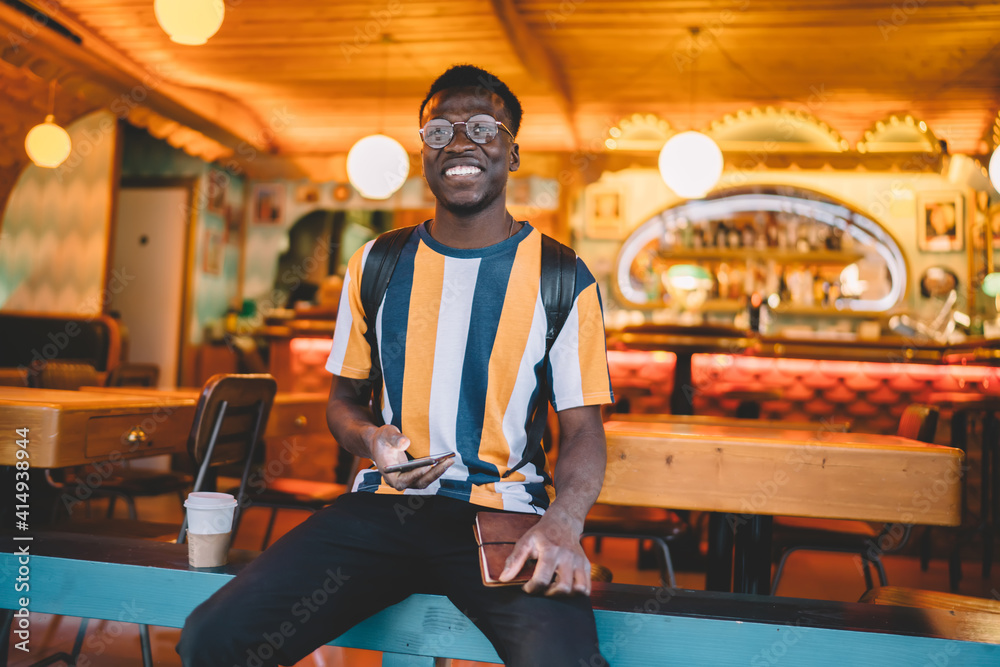 Portrait of cheerful male student in trendy spectacles for provide eyes correction holding cellphone technology and education textbook and smiling at camera, happy millennial with dark skin posing