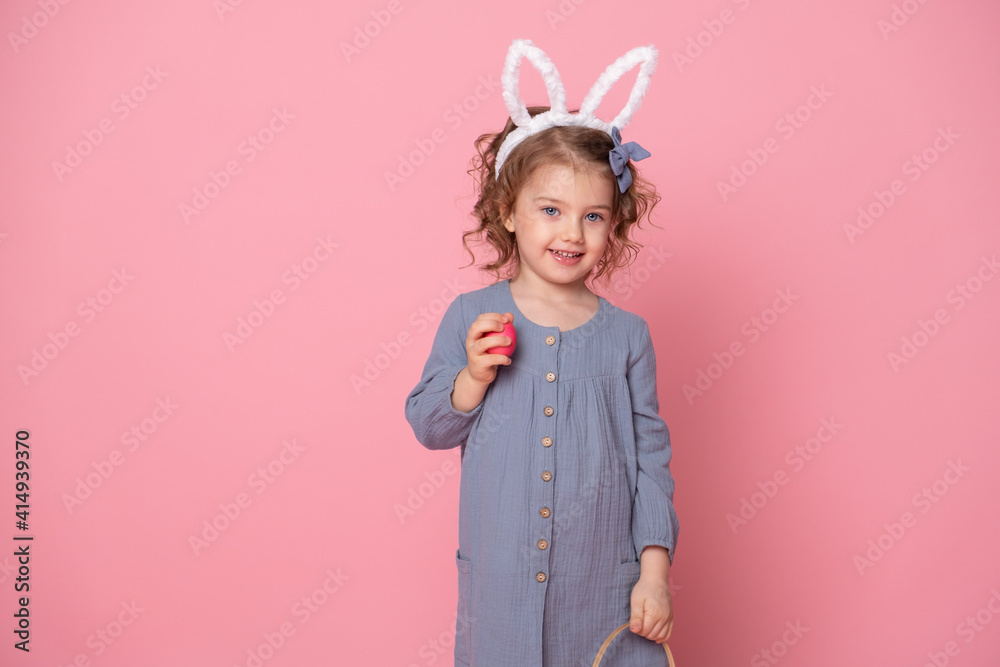 Funny child girl in Easter bunny ears holding basket with colorful eggs on pink background