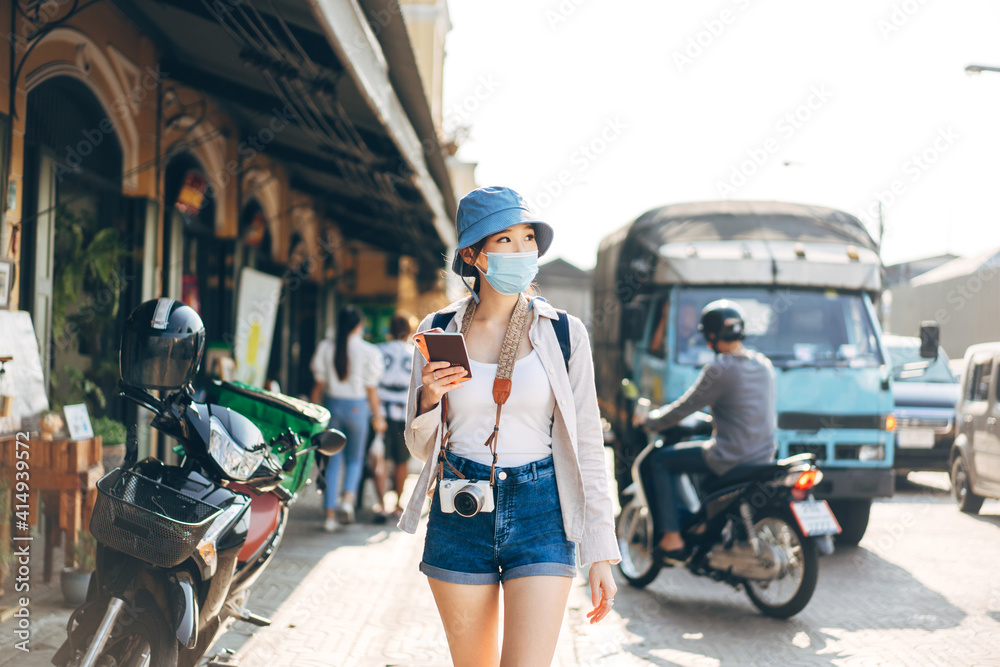 Walking young adult asian woman traveller wear face mask for covid-19 traveling in local city on summer day.
