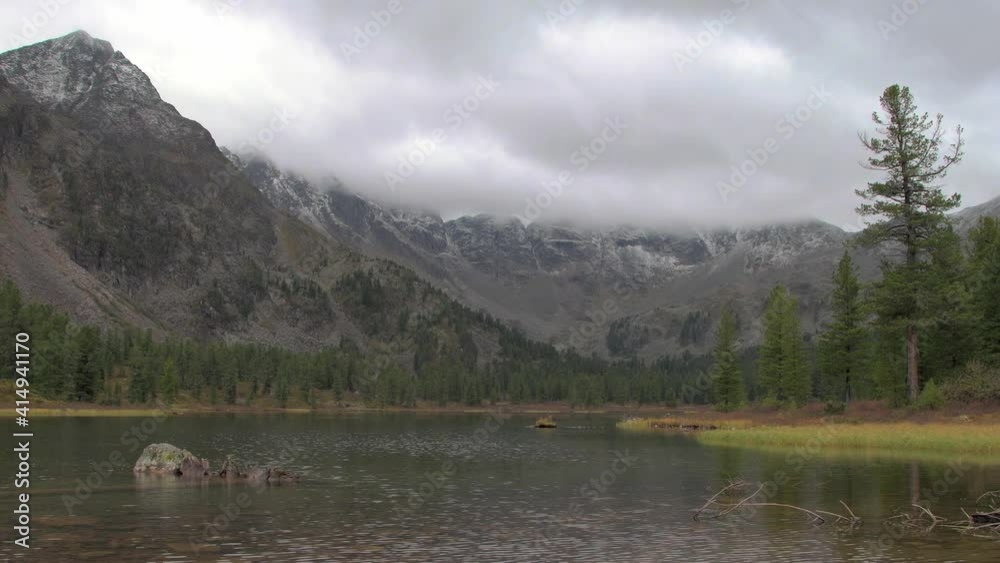 Above mountain range, sprinkled with snow, rain clouds float over it. Lake with stone is agitated by wind. Alone tree stands on grassy bank in autumn weather. Coniferous forest along river. Above mountain range, sprinkled with snow, rain clouds float over it. Lake with stone is agitated by wind. Alone tree stands on grassy bank in autumn weather. Coniferous forest along river.