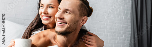 Smiling woman with cup hugging boyfriend at home, banner © LIGHTFIELD STUDIOS