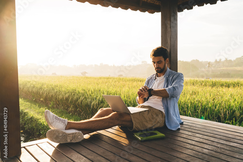 Side view of bearded guy in casual clothes checking wristband while browsing laptop while working on project remotely from wooden terrace among green area on summer day