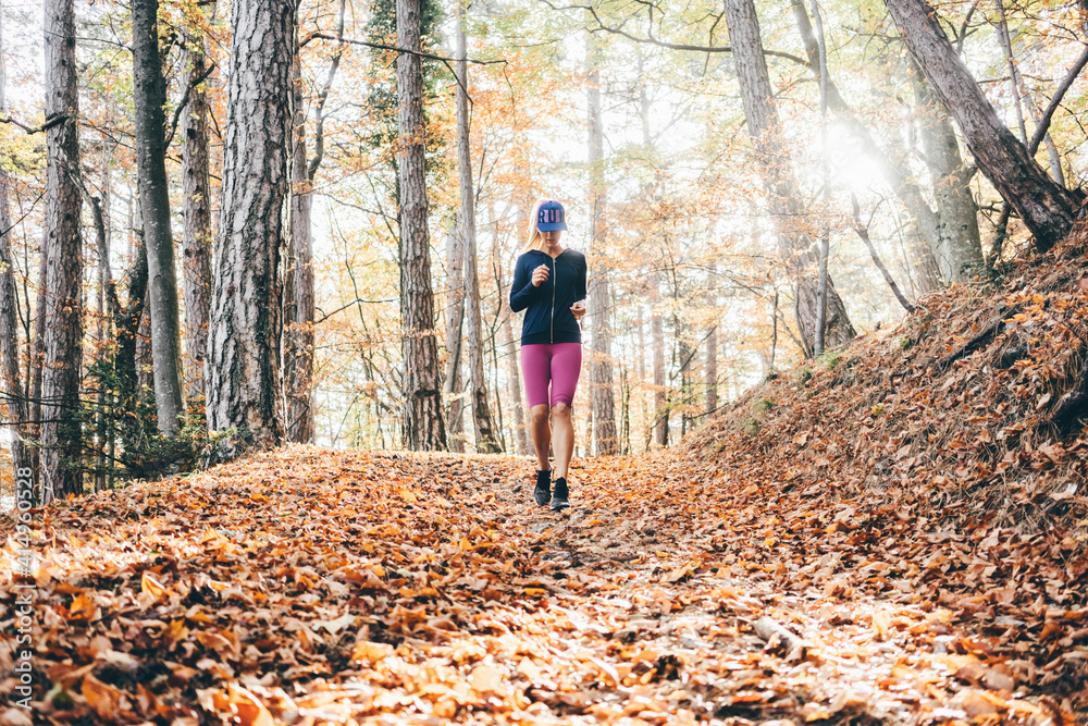 Smiling young woman with long blonde ponytail in tracksuit runs across picturesque autumn forest with old trees at bright back sunlight. Smiling young woman with long blonde ponytail in tracksuit runs across picturesque autumn forest with old trees at bright back sunlight.