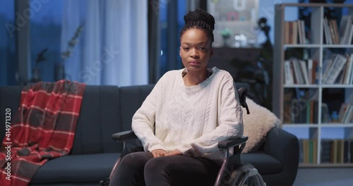 Portrait of serious afro-american disabled girl sitting in wheelchair posing for camera inside her house. Concept for illness, healthcare and medicine. photo