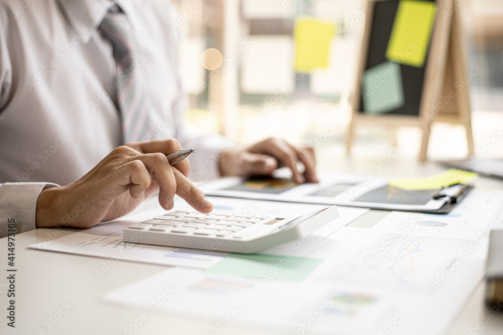 A business man is using a calculator to calculate company financial figures from earnings papers, a businessman sitting in his office where the company financial chart is placed. Financial concept. A business man is using a calculator to calculate company financial figures from earnings papers, a businessman sitting in his office where the company financial chart is placed. Financial concept.