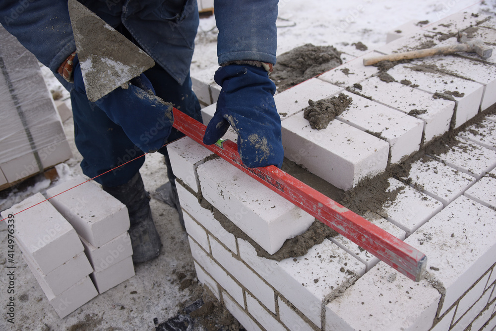 A builder in mittens puts gray bricks in cement in straight rows, makes a concrete wall.A bricklayer works on a construction site, laying concrete blocks, leveling a brick wall with a level.
