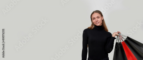 Studio shot of attractive young caucasian woman wearing black clothes smiling at camera, holding bunch of shopping bags while posing isolated over light gray background