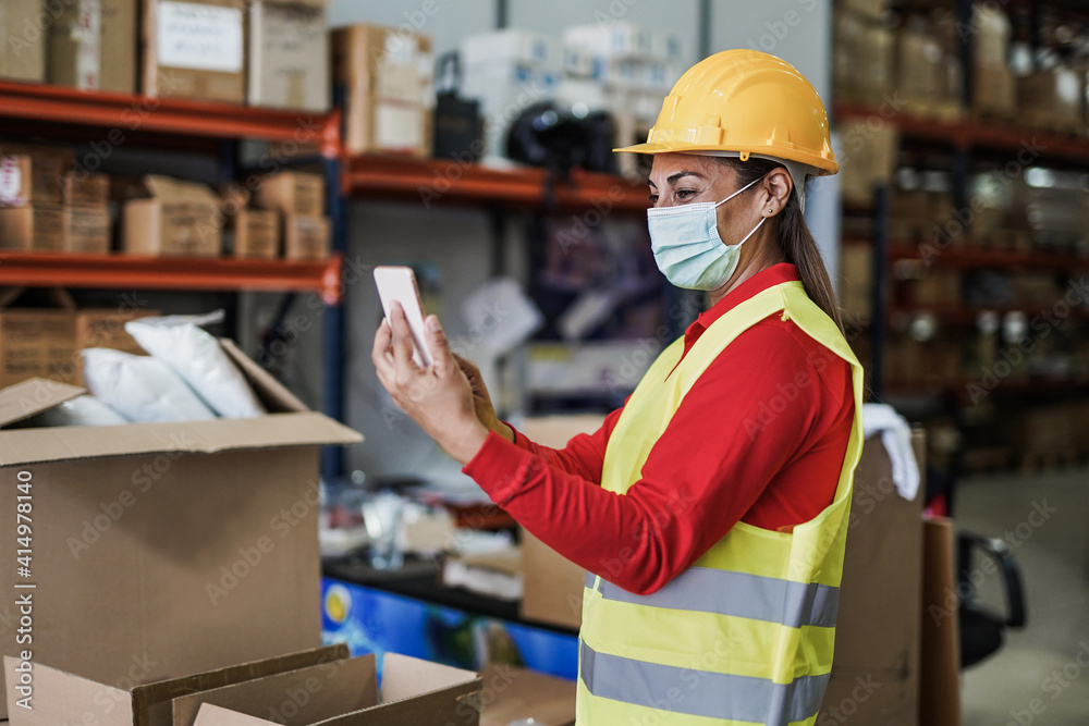 Mature latin woman with smartphone working inside warehouse while wearing safety face mask for coronavirus outbreak Mature latin woman with smartphone working inside warehouse while wearing safety face mask for coronavirus outbreak