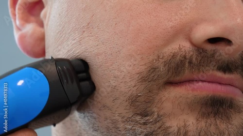 Male hand smoothly shaves stubble with shaving machine close-up. Man shaves his cheek with an electric safety razor. Shaving the beard on the face photo