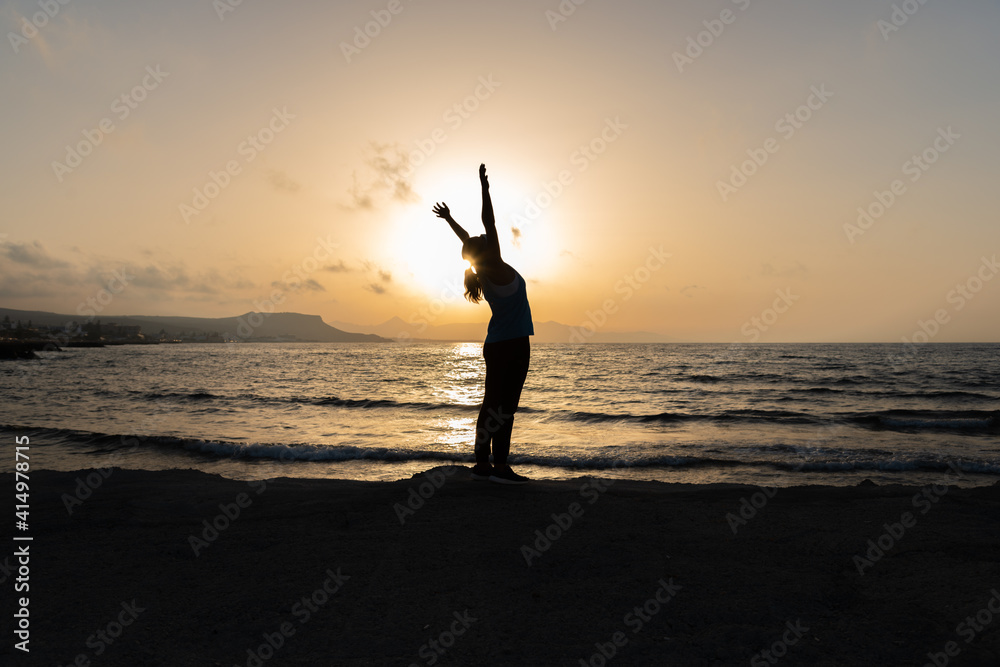 Silhouette of unrecognized young woman with protective surgical face mask performs yoga stretching exercises at the beach at dusk during coronavirus pandemic.