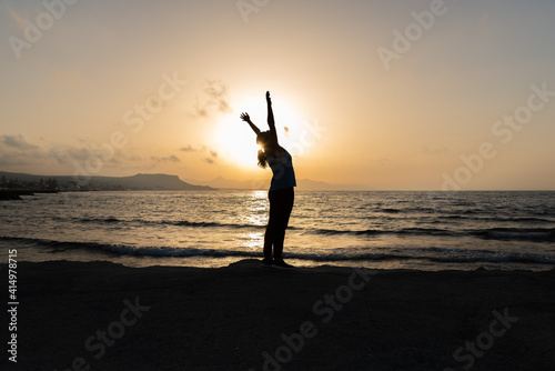 Obraz na płótnie Silhouette of unrecognized young woman with protective surgical face mask performs yoga stretching exercises at the beach at dusk during coronavirus pandemic. w salonie