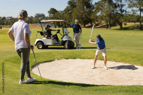 Four caucasian senior men and women playing golf watching one man preparing a shot from the bunker