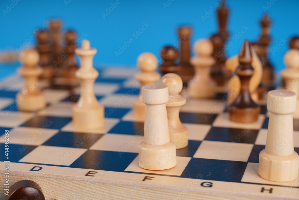 White and black wooden pieces on a chessboard. A chessboard set up during a game on a blue background. White and black wooden pieces on a chessboard. A chessboard set up during a game on a blue background.