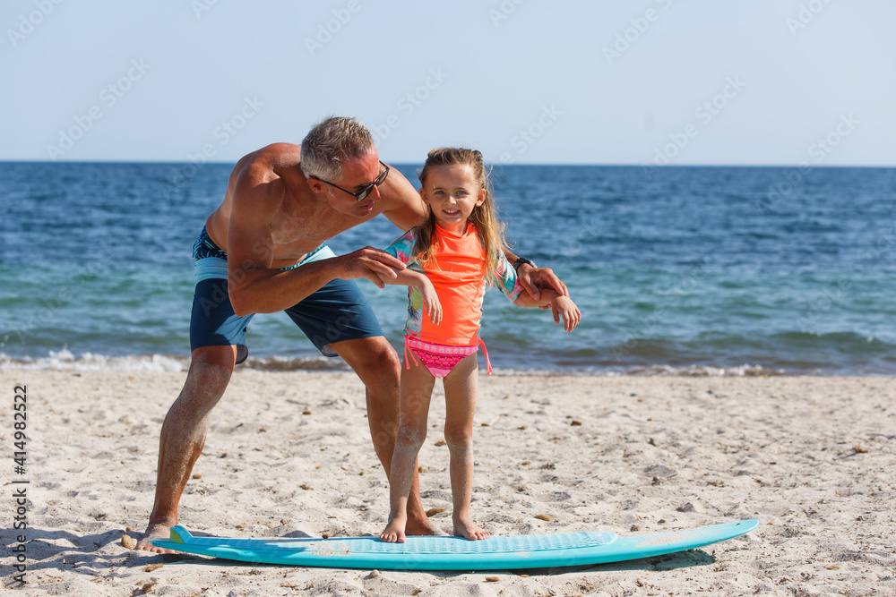 papà con occhiali da sole in costume da bagno insegna le basi del surf alla figlia in una spiaggia al mare.. papà con occhiali da sole in costume da bagno insegna le basi del surf alla figlia in una spiaggia al mare..