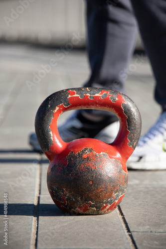 Close up shot of an old, very used red kettlebell on a paved flloor. Outdoor training session due to covid restrictions and gym closures in Italy. Trainer's feet in the background. photo