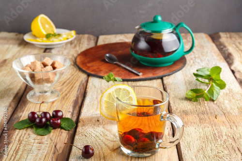 black tea in a glass cup with mint cherries and lemon on a wooden table next to fresh cherries and a teapot and lemon in a plate and mint leaves.