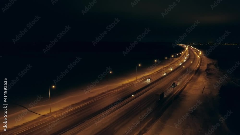 Aerial view of winter night highway with orange lanterns. Cars and trucks are driving along the highway. Near the highway is a village. There is a big city on the horizon. There is snow on the roadsid
