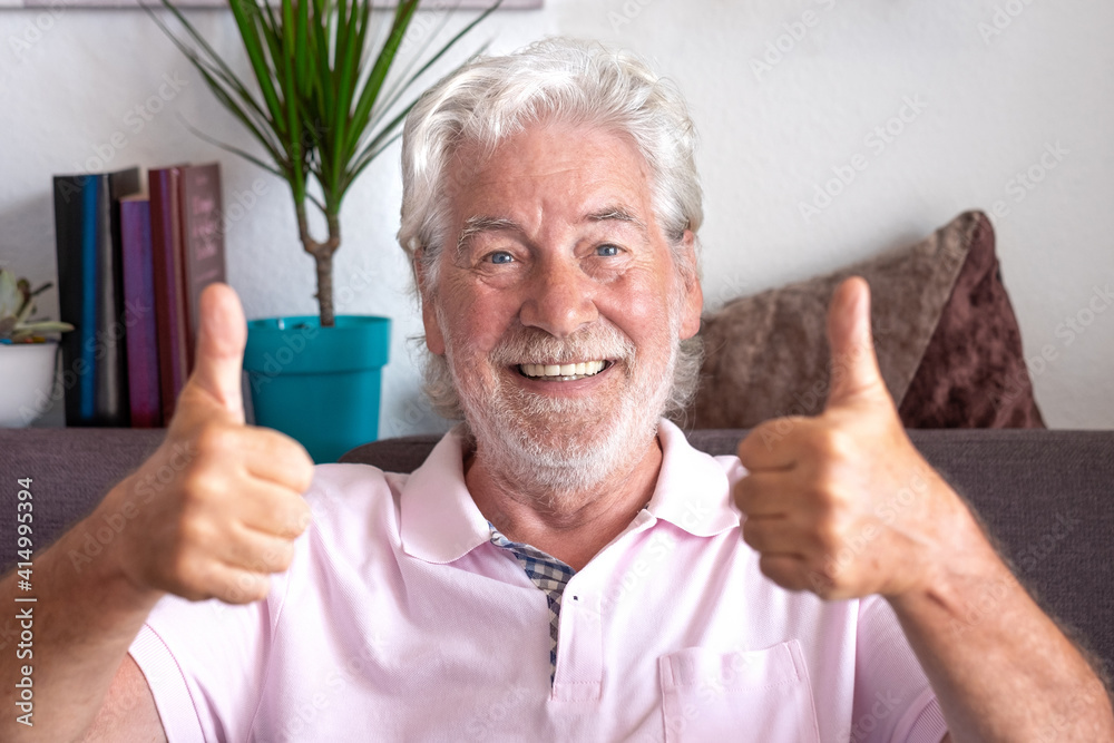 Cheerful white-haired senior man sitting at home on sofa making ok sign with hands, looking smiling into camera. Positive message from an elderly youthful grandfather Cheerful white-haired senior man sitting at home on sofa making ok sign with hands, looking smiling into camera. Positive message from an elderly youthful grandfather