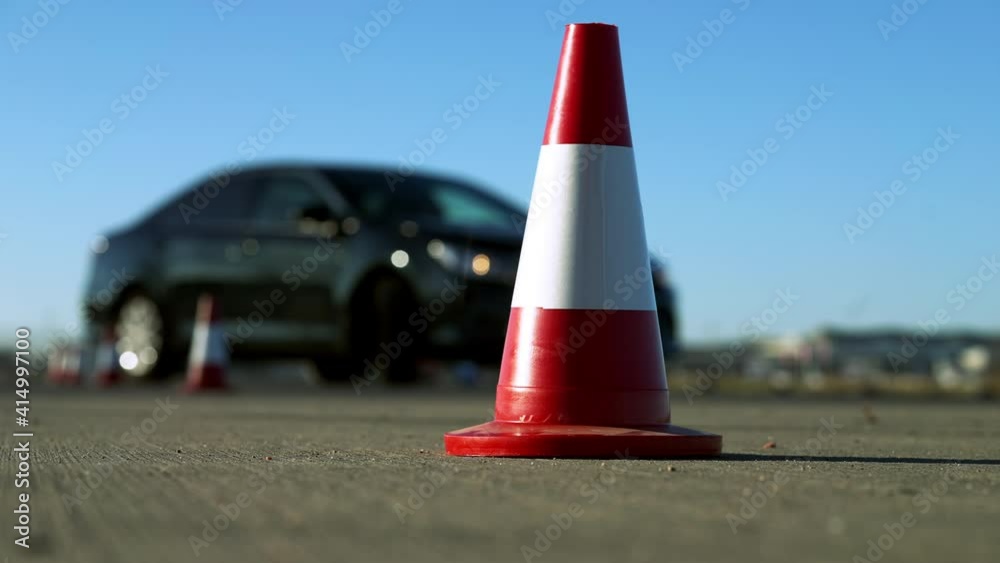 Close-up of red and white road cone with black car passing barriers. Unknown learner studying driving outdoors on sunny spring or auutmn day. Transportation and education concept.