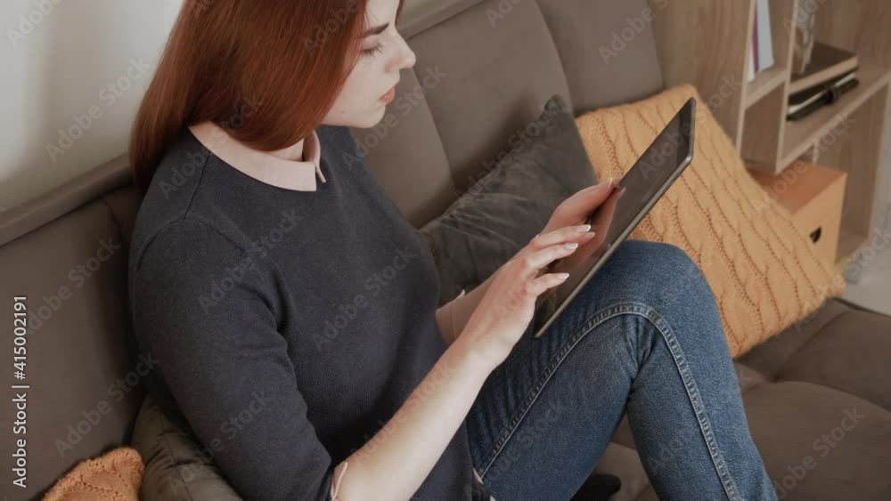 Happy young red hair female relaxing at home, playing game on modern tech device. Holding digital tablet, sit on sofa, enjoying surfing internet, study, work, shopping, using apps social media Happy young red hair female relaxing at home, playing game on modern tech device. Holding digital tablet, sit on sofa, enjoying surfing internet, study, work, shopping, using apps social media